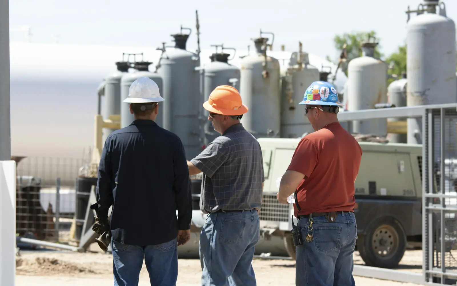 Three maintenance workers in hard hats seen from behind as they review equipment at a commercial facility yard